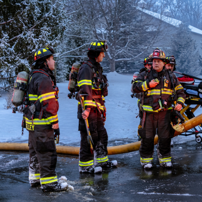 Firefighters in full gear stand near equipment and hoses at a snowy emergency scene, coordinating response operations.